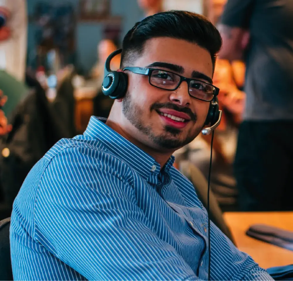 A man wearing glasses and a striped blue shirt smiles whilst sitting at a desk with a headset, suggesting he’s assisting customers with enquiries about modified 4x4 insurance in a call centre or customer service environment.