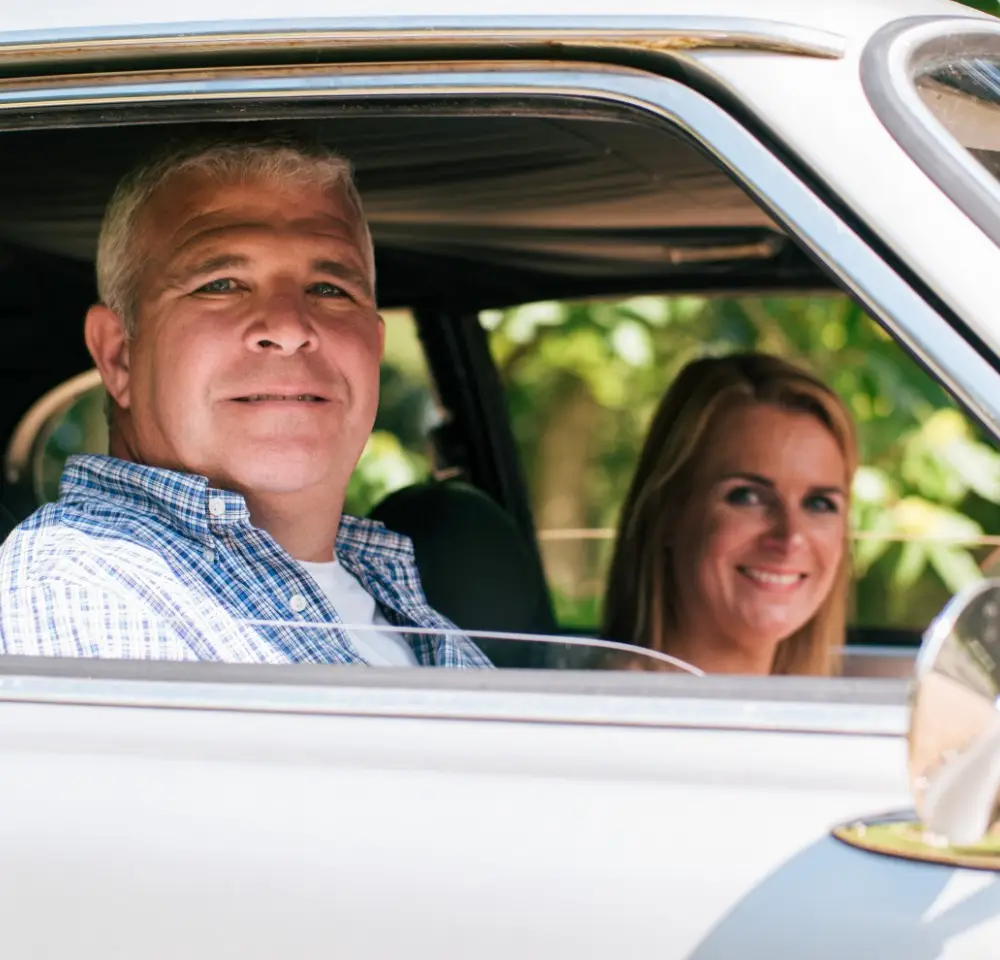 A man with grey hair and a checked shirt sits in the driver's seat of a car, smiling at the camera, while a woman with long blonde hair sits beside him, both enjoying their drive—protected by reliable modified 4x4 insurance—with greenery outside the window.