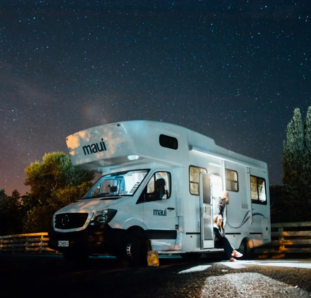 A person stands at the open door of a lit-up campervan labelled “maui” beneath a clear, starry night sky, with trees and a fence in the background—reminding travellers of the importance of Motorhome Insurance for peace of mind on every journey.