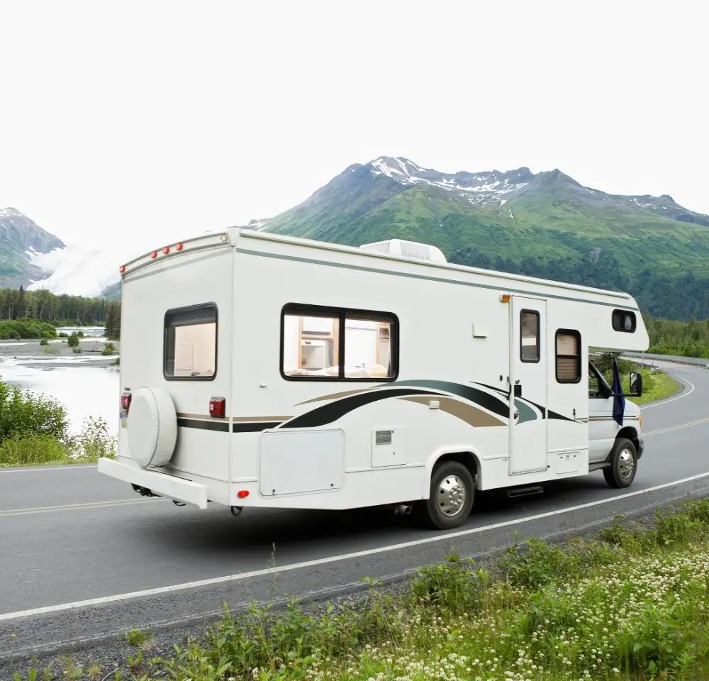 A white motorhome with green and black stripes is parked on a scenic road beside a river, with green mountains and snow-capped peaks in the background—an ideal setting to consider the benefits of Motorhome Insurance.