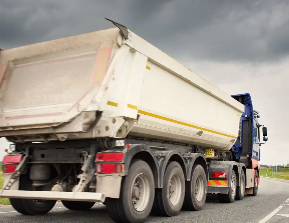 A large dumper lorry with a white trailer, ideal for those seeking Tipper Insurance, drives on a road under a cloudy, dark sky. Captured from a low rear angle, the lorry’s multiple wheels highlight its size and industrial purpose.