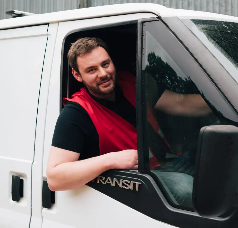 A man wearing a red waistcoat and black shirt sits in the driver’s seat of a white van, leaning out of the open window and smiling at the camera—perfect for showcasing any driver van insurance for business use.
