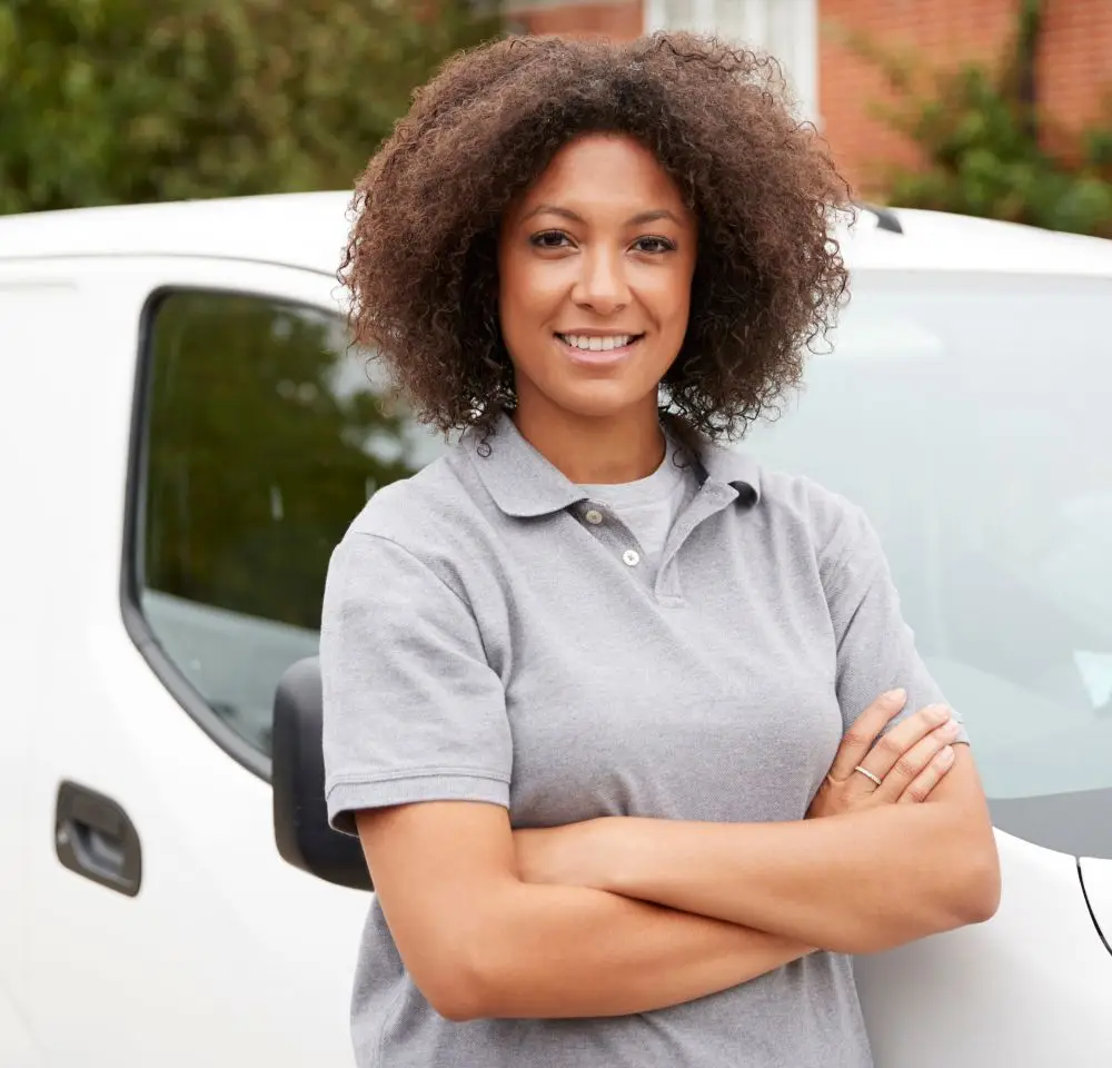 A woman with curly hair, wearing a grey polo shirt, stands smiling with arms crossed in front of a white van and a brick building—perfectly representing the confidence that comes with reliable Business Van Insurance.