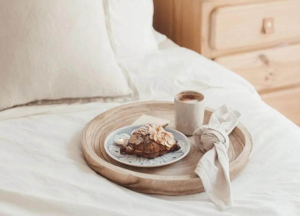 A wooden tray on a neatly made bed holds a cup of coffee, a pastry topped with sliced almonds on a plate, and a cloth serviette, creating a cosy breakfast-in-bed scene perfect for showcasing your Bed and Breakfast Insurance offerings.