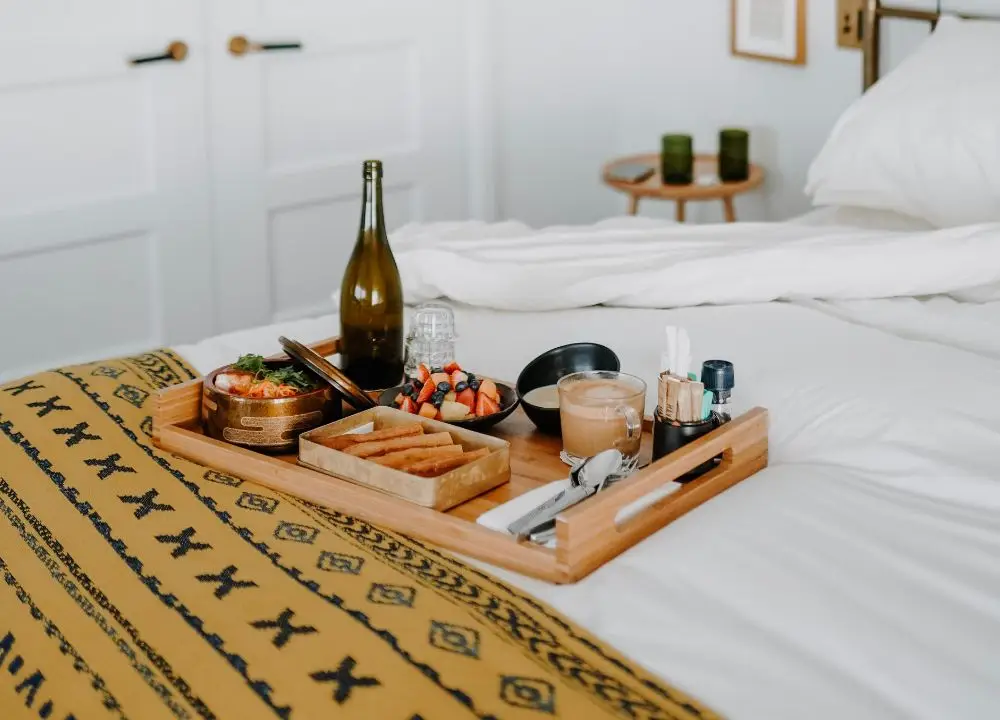 A wooden tray with breakfast food, coffee, a bottle, and various dishes sits on a bed with white sheets and a yellow patterned blanket—capturing the welcoming spirit of Bed and Breakfast Insurance. The background includes a small round table and a white wall.