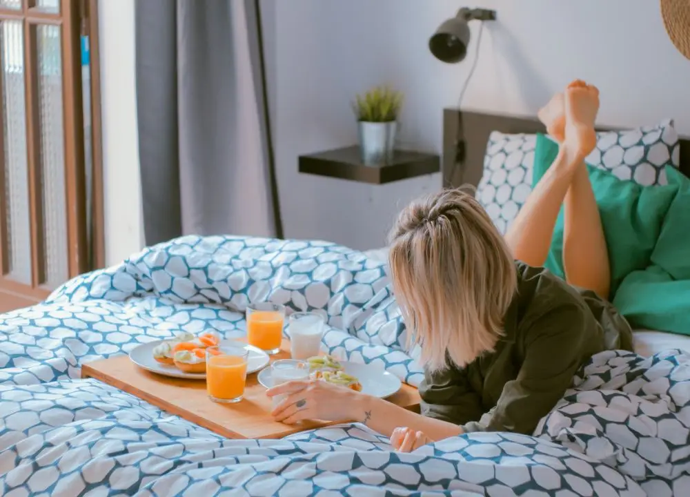A woman with blonde hair lies on a bed with geometric-patterned bedding, facing away from the camera. She enjoys a breakfast tray in a room with cosy, modern decor—an inviting scene that highlights the comfort Bed and Breakfast Insurance helps protect.