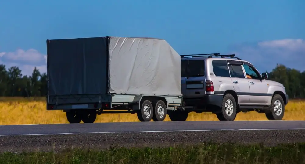 A silver SUV drives down a rural road, towing a covered car transporter with two axles. The trailer, wrapped in a grey tarpaulin, highlights the need for trailer insurance as it passes fields, trees, and blue sky in the background.