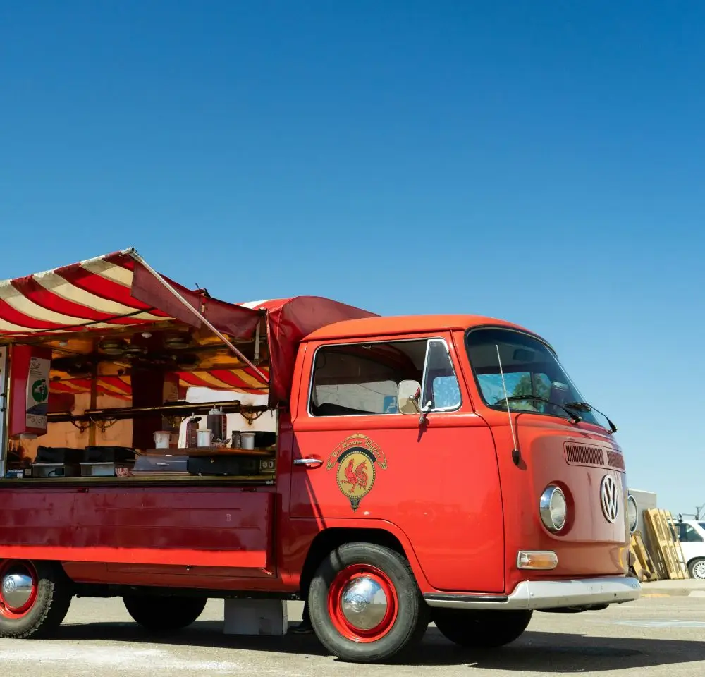 A vintage red Volkswagen van, covered by Classic Commercial Vehicle Insurance, is converted into a food truck and parked under a clear blue sky. The side awning is open, revealing the kitchen and serving area inside.
