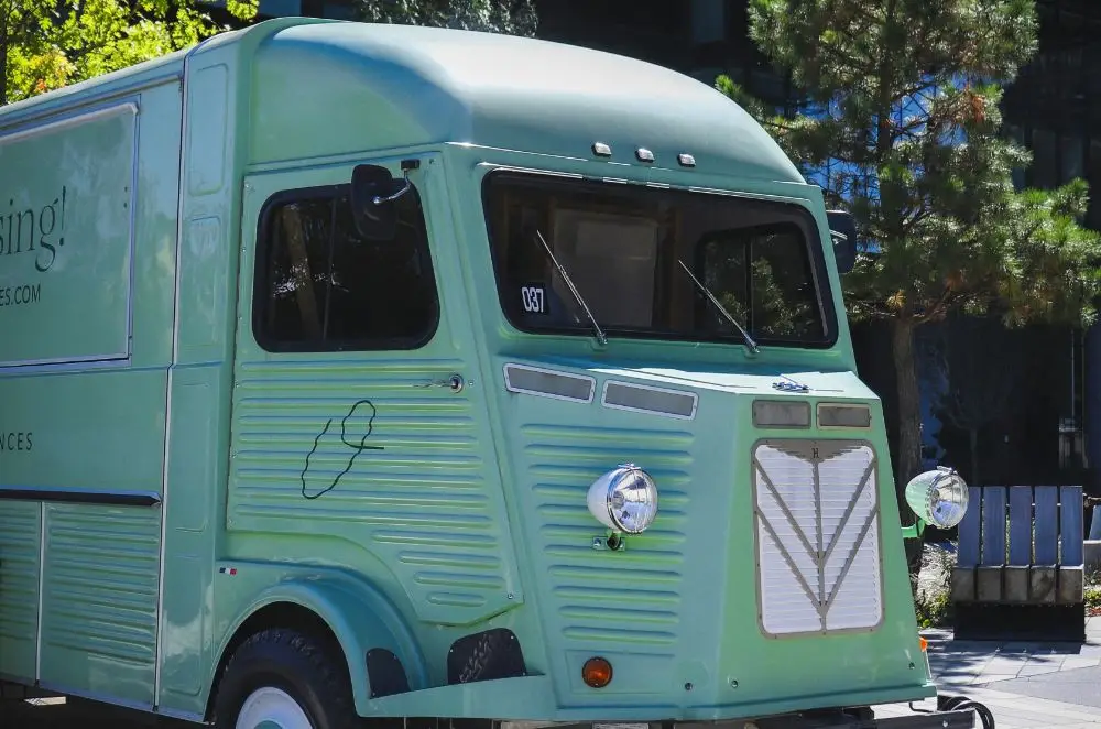 A vintage green food van with a retro design, covered by Classic Commercial Vehicle Insurance, is parked outdoors near trees and benches on a sunny day.