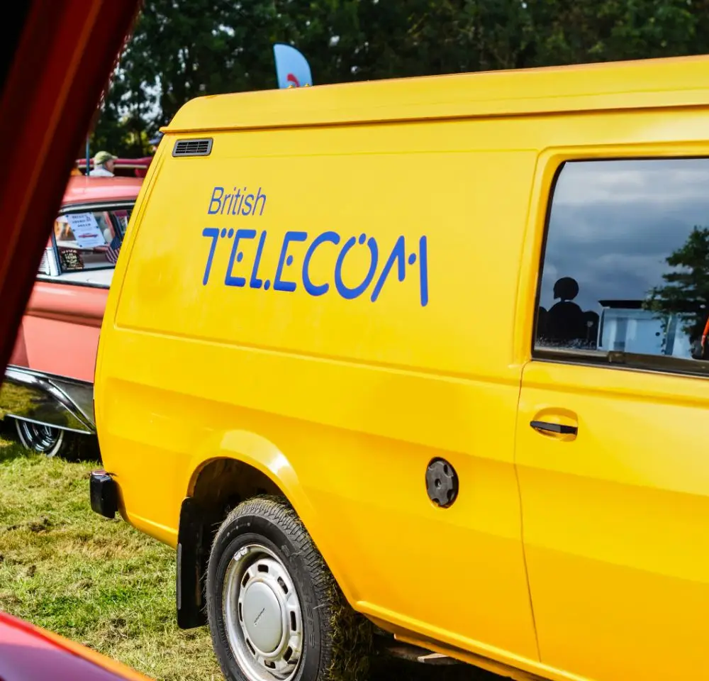 A bright yellow van with British Telecom written on the side in blue letters is parked on grass, with part of another vehicle beside it. Perfect for Classic Commercial Vehicle Insurance enthusiasts, trees and a cloudy sky form the background.