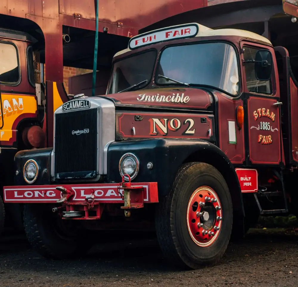 A vintage red and black Scammell lorry labelled “Invincible No 2” and “Steam Fair” is parked, featuring chrome details, red wheels, and signs that read “Fun Fair” and “Long Load”—perfect for Classic Commercial Vehicle Insurance enthusiasts.