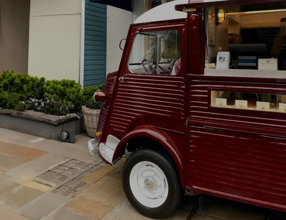 A vintage red food van parked on a stone street, with a display of small packaged goods in the window and green shrubs in the background.