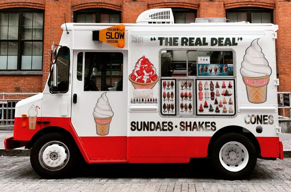A red and white ice cream van, covered in images of cones, sundaes, and milkshakes, sits on a city street. With its menu window and THE REAL DEAL slogan, it’s a perfect example of why Classic Van Insurance matters for beloved vehicles like this.