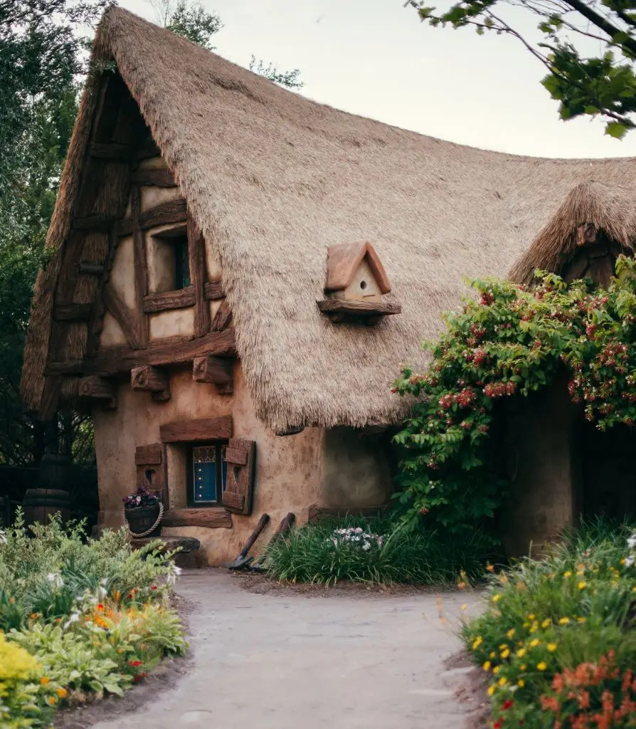 A charming cottage with a thatched roof, rustic wooden beams, and a small blue door sits amidst lush greenery and flowers—an ideal retreat where Cob House Insurance offers peace of mind beneath the soft, overcast sky.