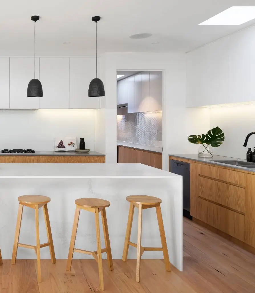 Modern kitchen with white worktops, wooden cupboards, three bar stools, black pendant lights, and a sleek black tap. A large green leaf in a vase adds charm—ideal for those seeking stylish spaces and peace of mind with rental property insurance.
