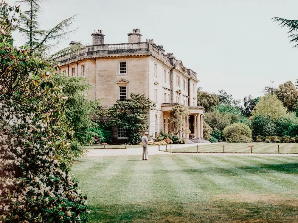 A person stands on a manicured lawn in front of a large, elegant historic mansion—an ideal property for High Value Home Insurance—surrounded by trees and lush greenery on a bright day.