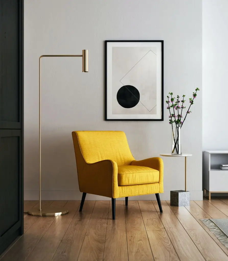 A modern living room with a yellow armchair, gold floor lamp, round side table with a vase of branches, and abstract art on light walls—an inviting space where High Value Home Insurance helps protect your elegant minimalist décor.