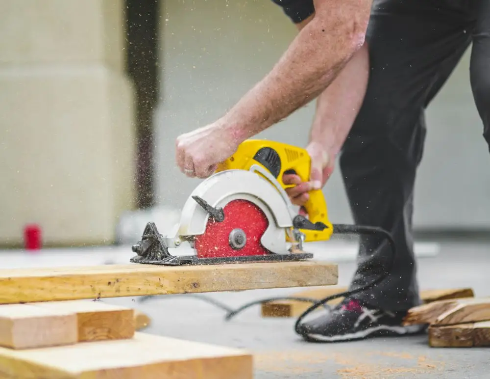 A person uses a yellow circular saw to cut a wooden plank outdoors, sawdust flying in the air. Dressed in black trousers and trainers, they're tackling a DIY project—reminding us how renovation insurance can protect your efforts.