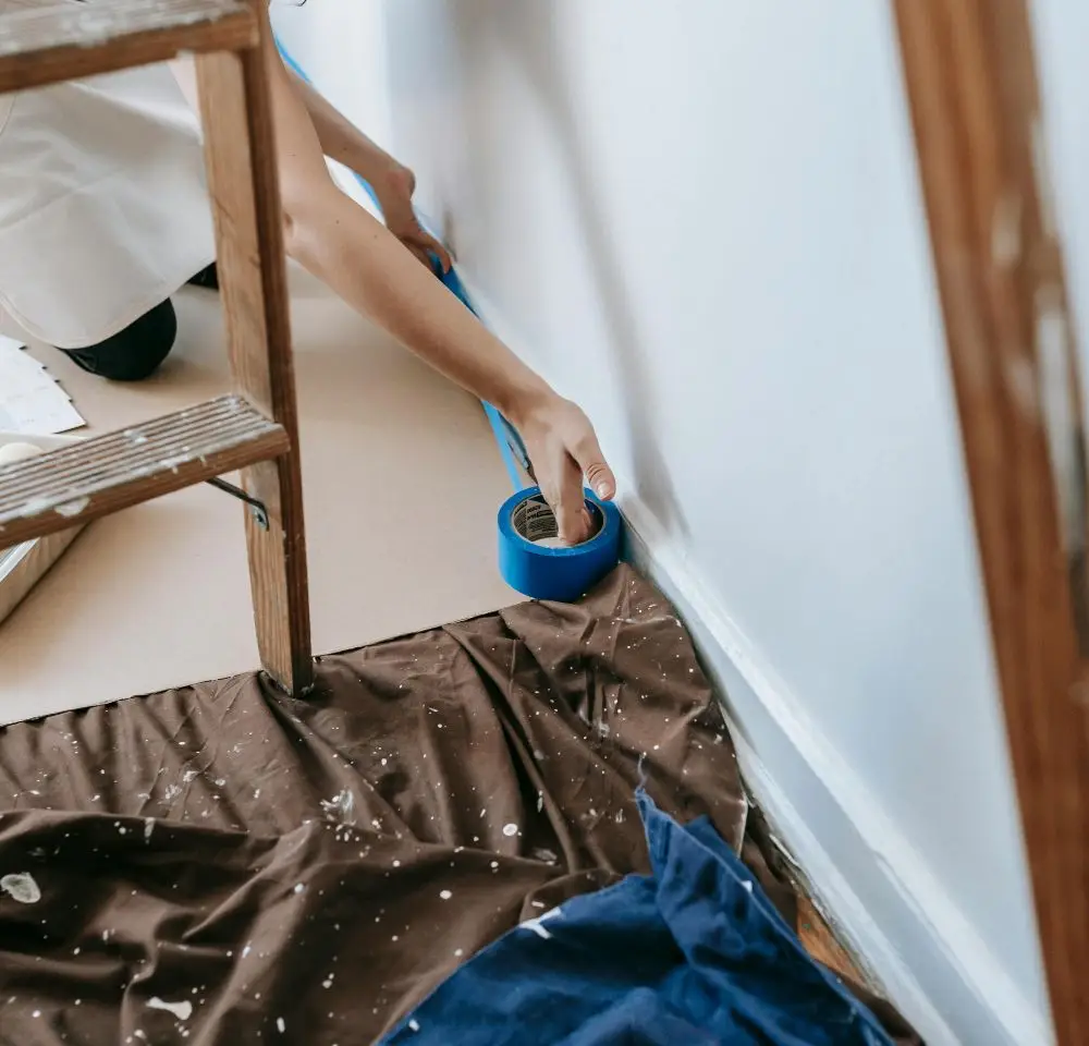 A person kneels on the floor, applying blue painter’s tape along the skirting board of a wall, with a brown dust sheet spread out and a wooden ladder nearby—an ideal scene to consider Home Renovation Insurance for added peace of mind.