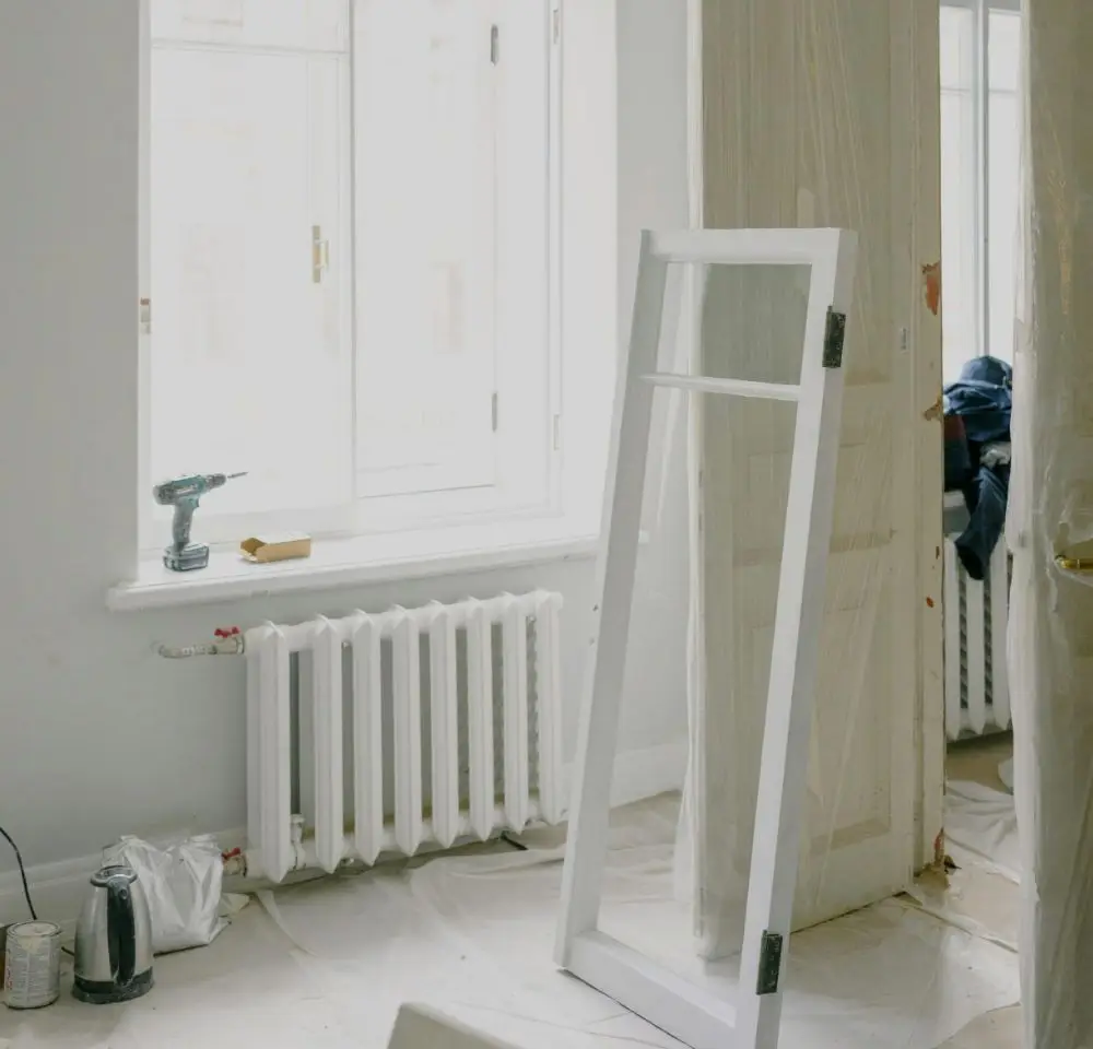 A bright room undergoing renovation with a window, radiator, detached door or window frame, a drill on the windowsill, and protective plastic sheets covering parts of the space—an ideal scene to consider Home Renovation Insurance for added peace of mind.