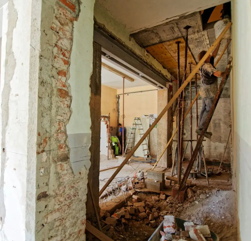 A construction worker stands on wooden scaffolding inside a partially demolished building, surrounded by debris, exposed brickwork, and tools—reminding homeowners of the importance of home renovation insurance. Another person works in the background near a ladder.