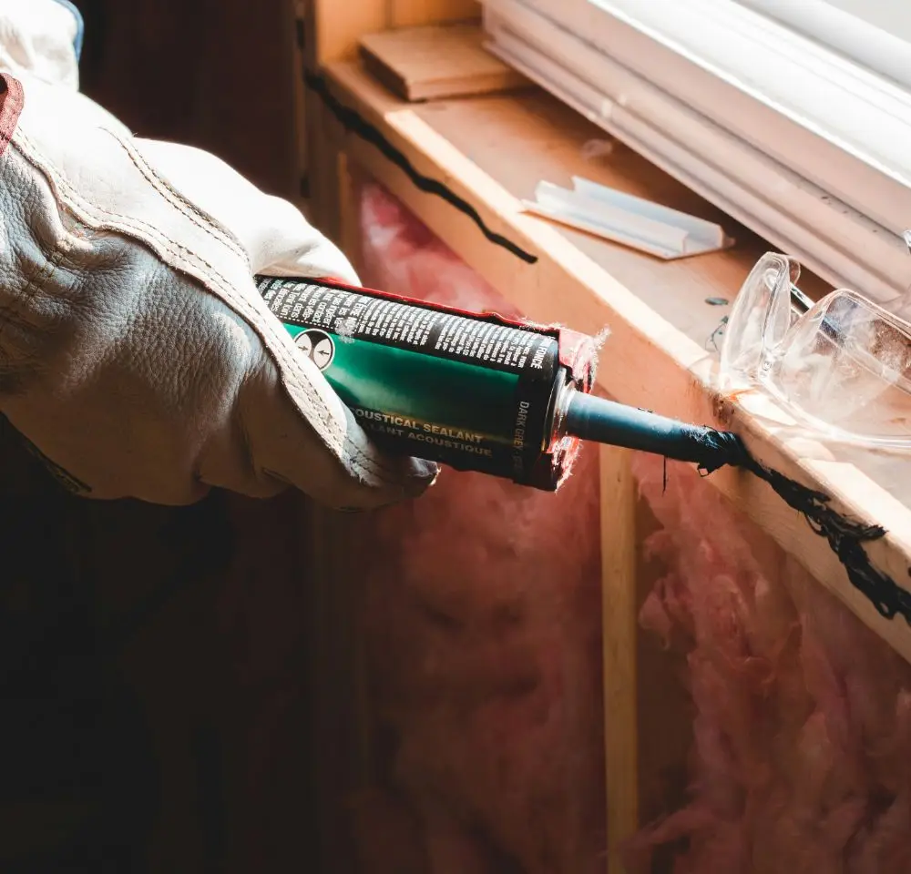 A person wearing work gloves uses a caulking gun to seal a window frame, with insulation and protective goggles nearby—an essential step for those considering home renovation insurance.