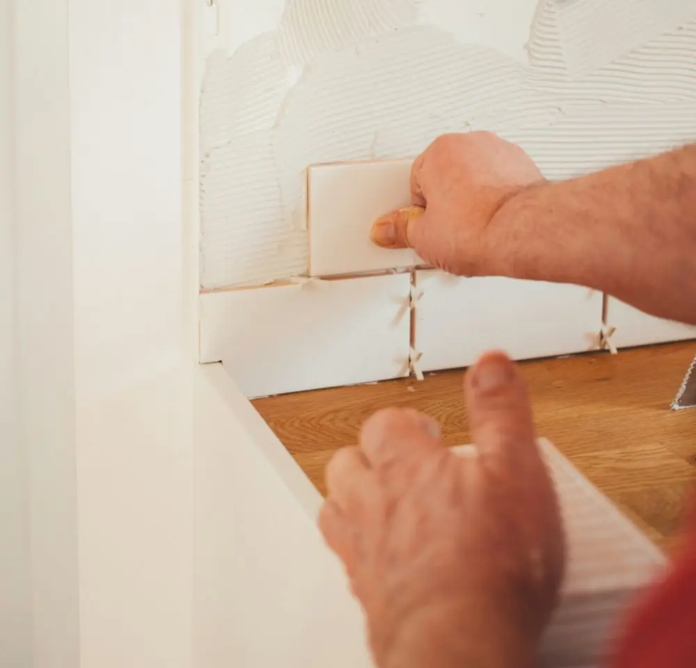 A person installs a white rectangular tile on a kitchen splashback, pressing it onto adhesive with spacers between tiles—an important step in any refurbishment where Home Renovation Insurance can help protect your investment.