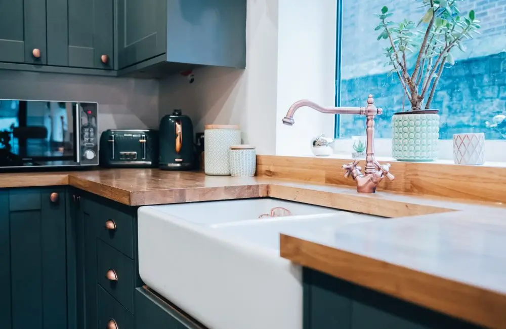 Modern kitchen with green cupboards, wooden worktops, a butler sink, copper tap, and small appliances—perfect for any home. Landlord insurance can help protect such beautiful spaces filled with natural light from unexpected damages.