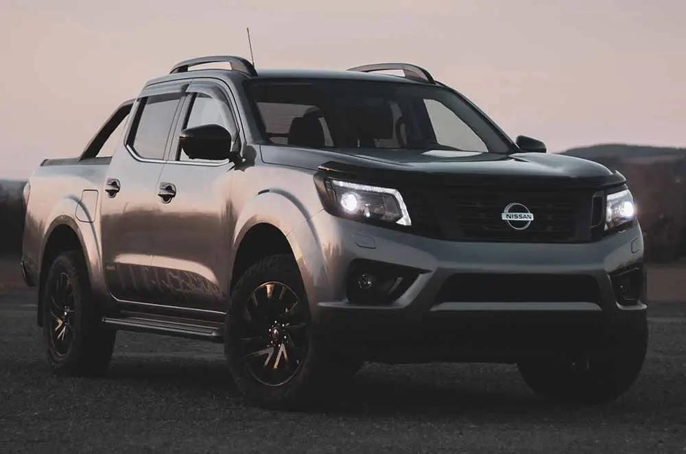 A silver Nissan pick-up truck parked outdoors at dusk, with dark alloy wheels and roof rails visible, against a muted, mountainous background.