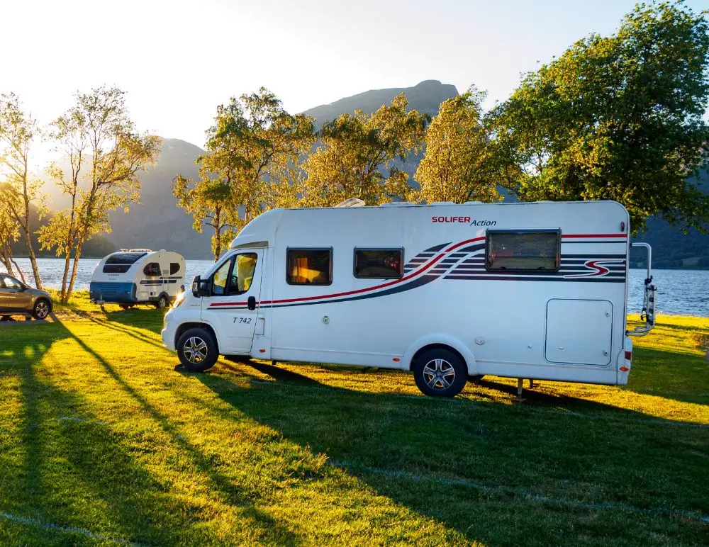 A white camper van parked on grassy ground near a lake, with trees and mountains in the background. Another camper and a car are visible nearby in the sunlight—a perfect scene to remind you of the importance of Motorhome Insurance.