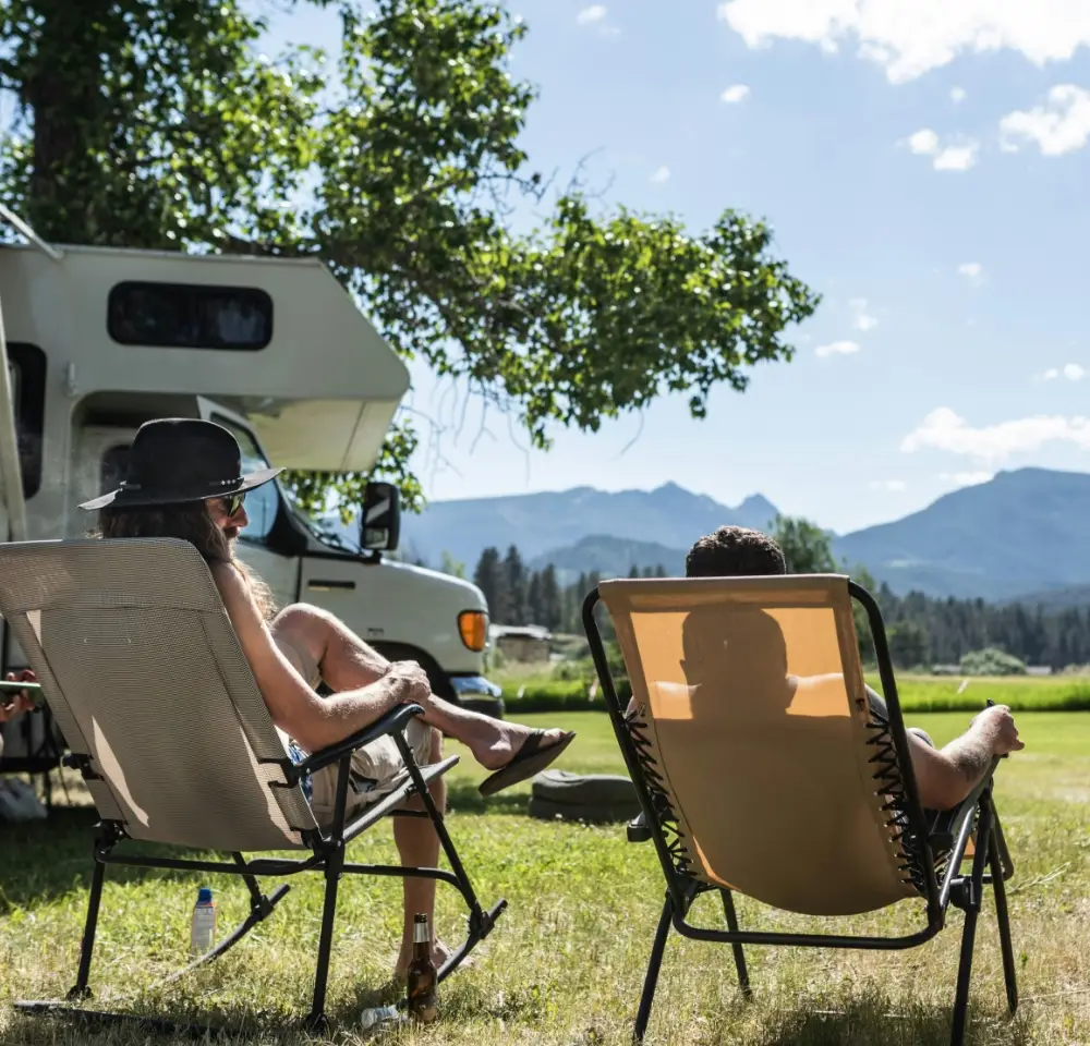 Two people relax in loungers on grassy ground beside their campervan, enjoying a sunny day with mountains and trees in the background. A bottle sits nearby, setting the scene for a peaceful camping trip—ideal for those with Motorhome Insurance.