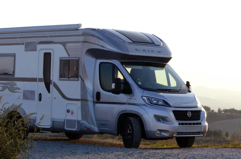 A silver motorhome with modern design features is parked on a gravel road, with hills and a clear sky in the background—a perfect scene to highlight the importance of motorhome insurance.