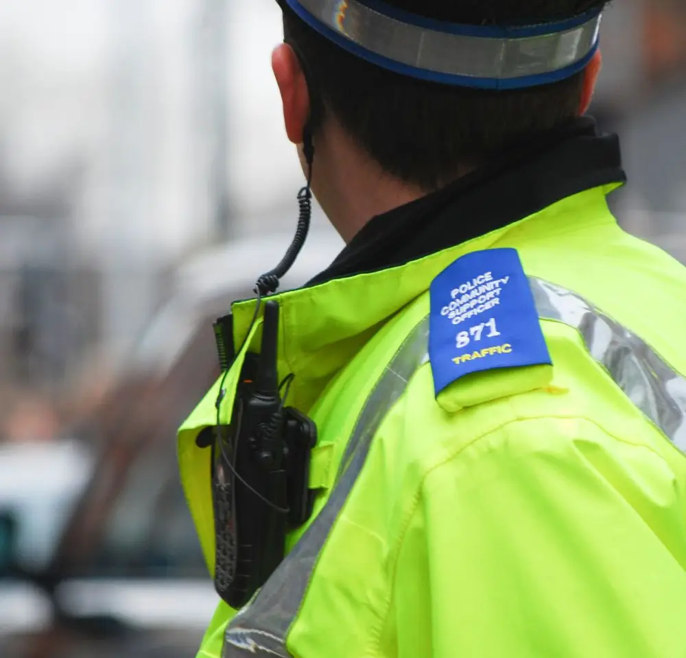 A Police Officer in a bright yellow jacket with a radio stands outdoors, seen from behind. The blue shoulder badge reads “POLICE COMMUNITY SUPPORT OFFICER 871 TRAFFIC,” highlighting their role in traffic safety and car insurance checks.
