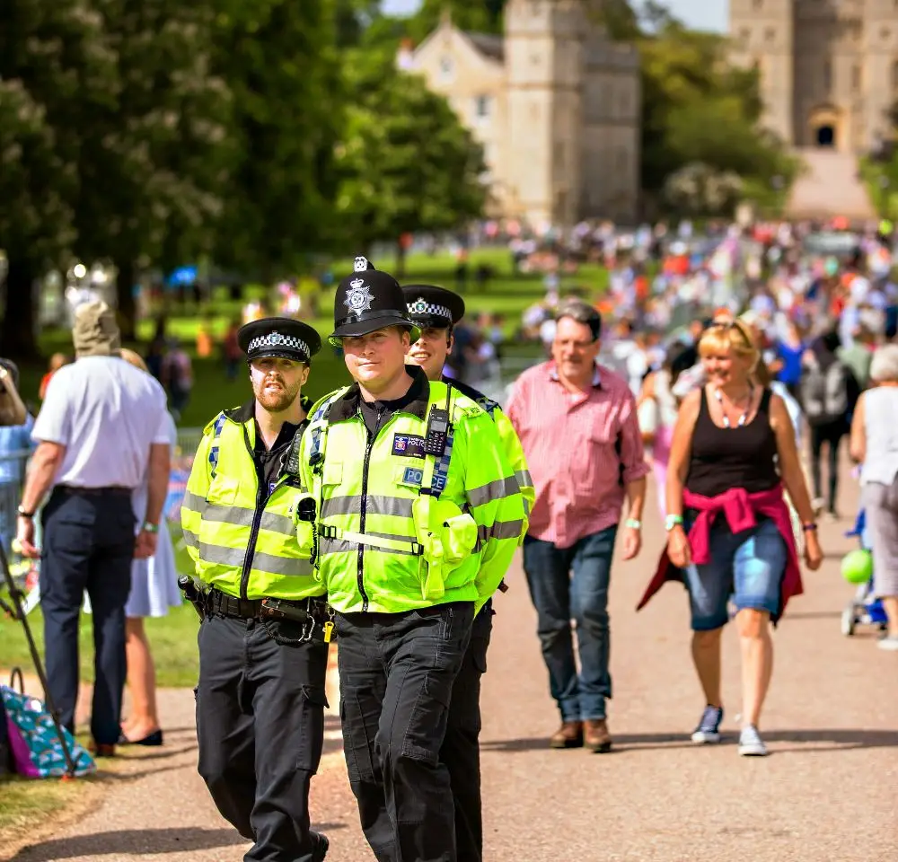 Three police officers in high-visibility jackets walk along a path in a busy park on a sunny day, surrounded by people, with a large historic building in the background.