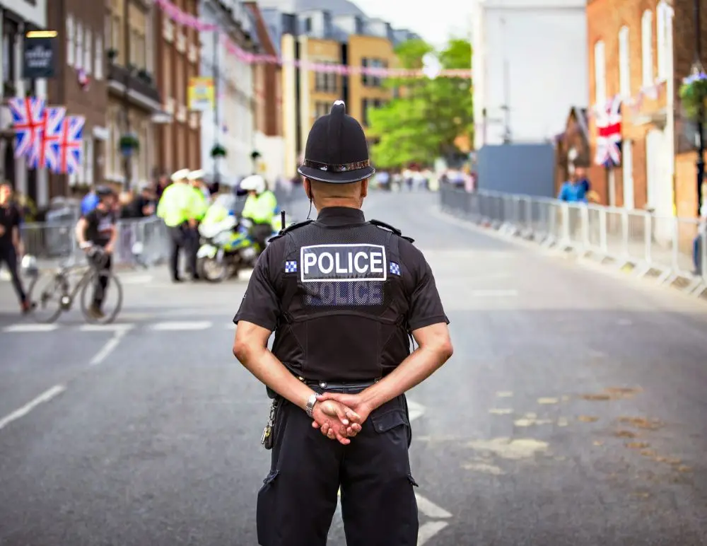 A police officer stands in the middle of a city street with hands behind his back, facing away. There are people, bicycles, other officers, and Union Jack flags visible—no sign of car insurance checks here.