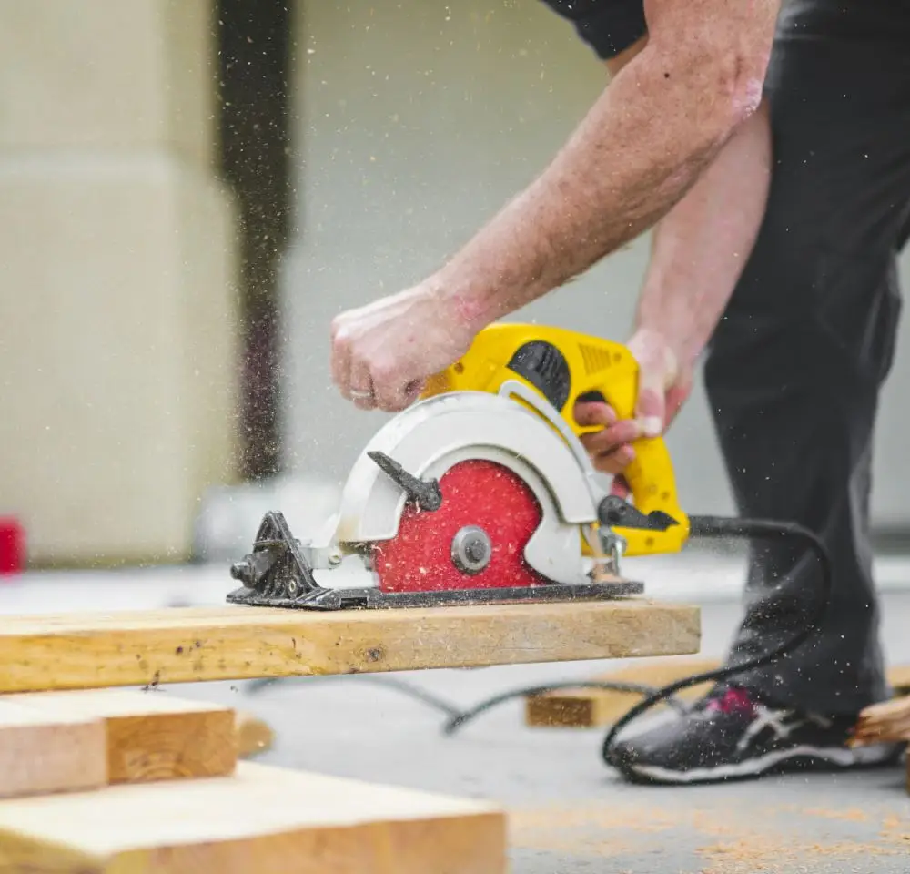 A person uses a yellow electric circular saw to cut a wooden plank outdoors, sawdust flying in the air. Their hands and arms are visible as they work safely—a key consideration for anyone with Self Build Home Insurance.