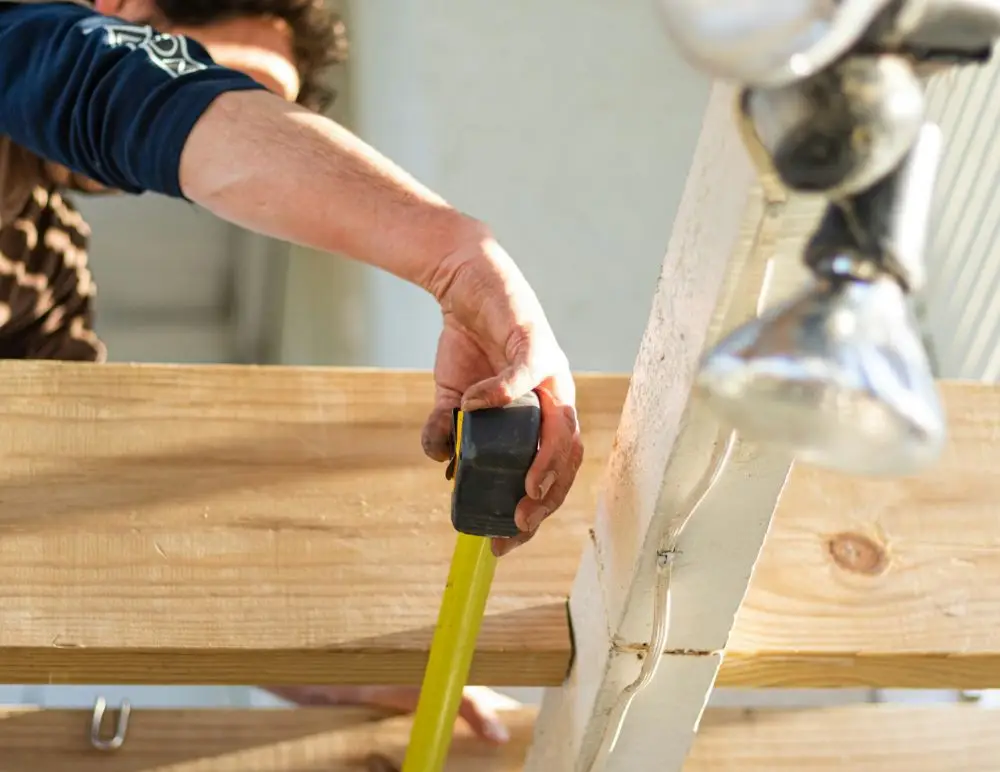 A person measures a wooden beam with a yellow tape measure, holding it in place with one hand. Bright sunlight illuminates the scene, highlighting the wood and the person's arm—an essential step for any self build home insurance project.