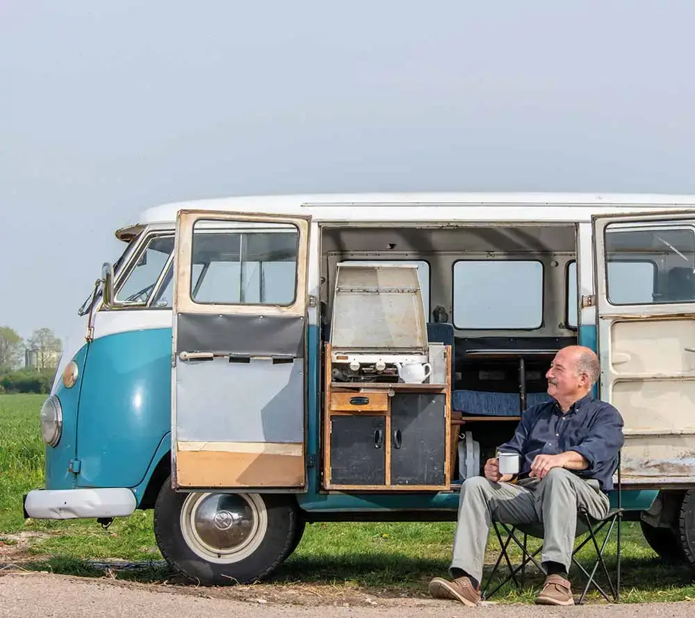 An older man sits on a folding chair next to a classic blue and white camper van, enjoying his mug and the view by a grassy field. With Temporary Campervan Insurance, he’s ready for carefree adventures under the clear sky.