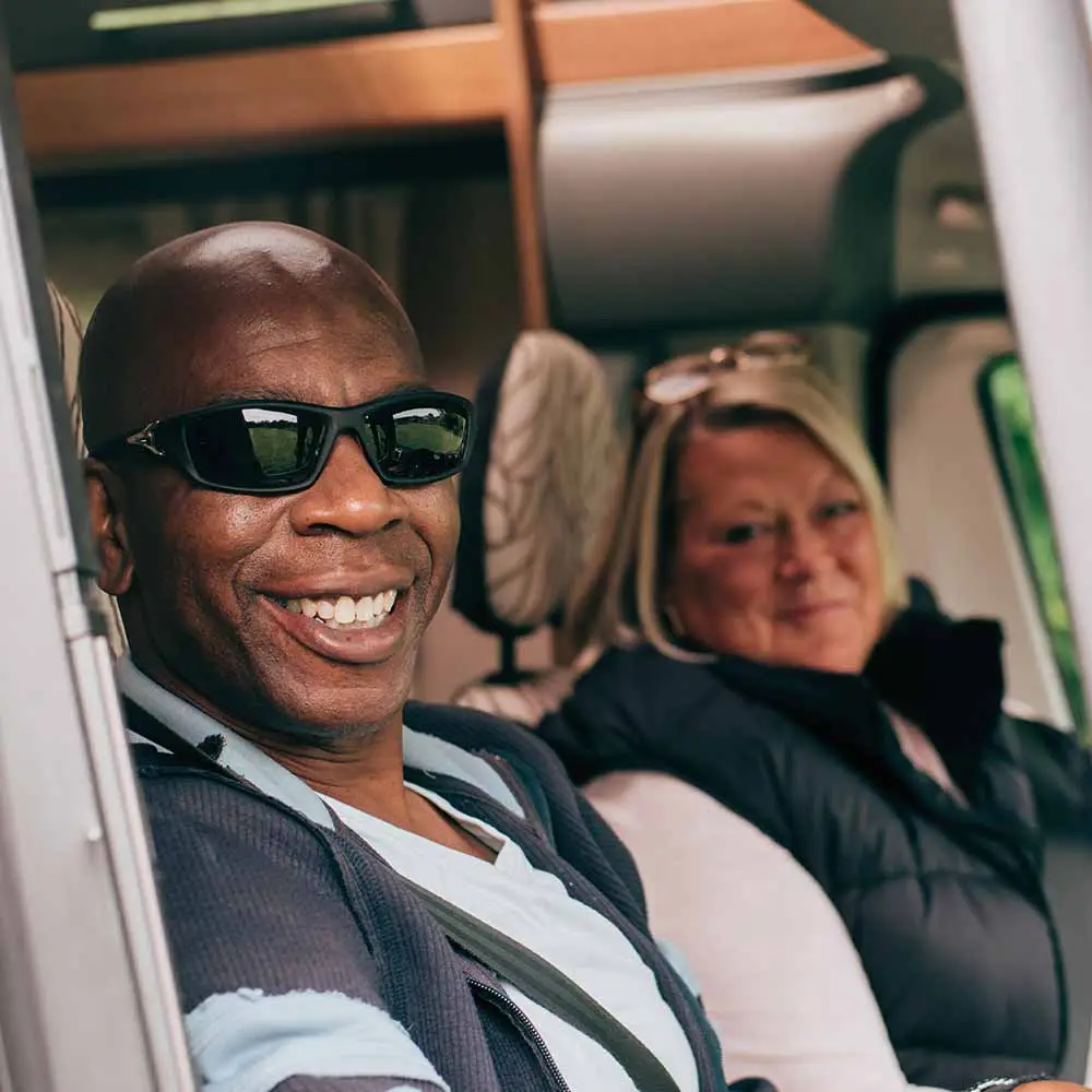 A man wearing sunglasses smiles at the camera from the driver’s seat of a vehicle, possibly enjoying a trip with Temporary Motorhome Insurance. Beside him, a woman looks towards the camera and smiles softly.