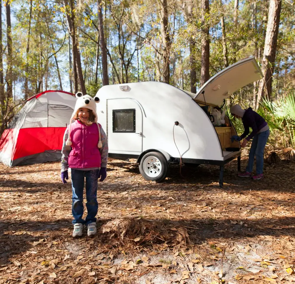 A young girl in a pink waistcoat and animal-ear hat stands in front of a small caravan and tent at a wooded campsite, while another person, perhaps thinking about caravan insurance, prepares something at the caravan’s open hatch.