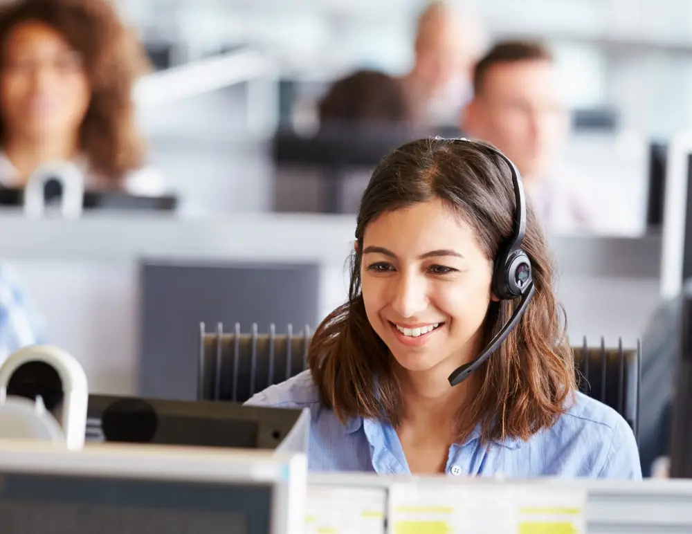 A woman wearing a headset smiles whilst sitting at her desk in an office, appearing to assist customers as part of a call centre team with blurred colleagues in the background.