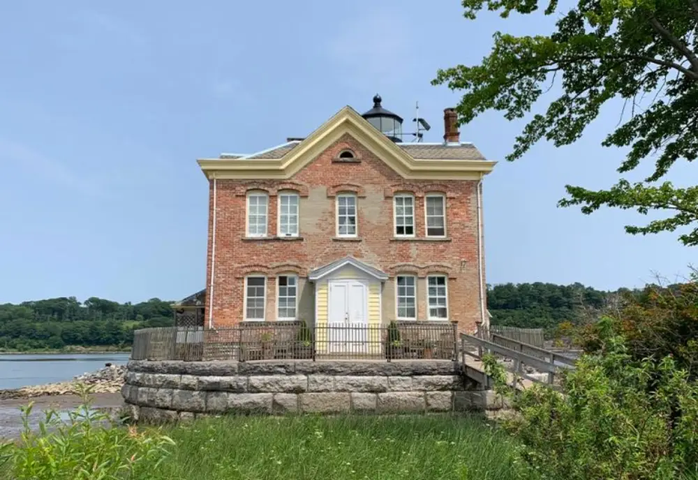 A historic brick lighthouse with a yellow door and white trim sits on a stone foundation by the water, surrounded by greenery—an ideal candidate for Converted Property Insurance under the clear sky.