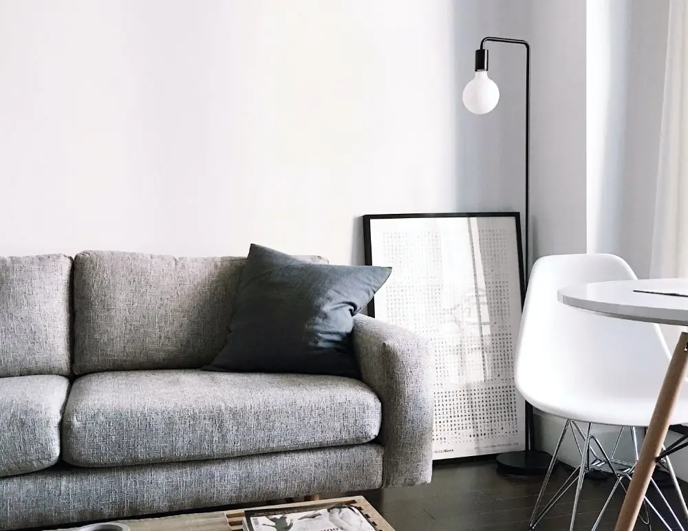 A modern living room with a light grey sofa and dark cushion, white framed artwork leaning against the wall, a round white table with white chairs, and a black floor lamp—perfect for showcasing the comfort offered by HMO Landlord Insurance.
