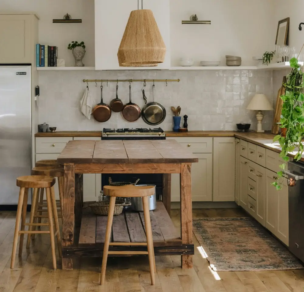 A cosy, rustic kitchen with light cabinetry and a wooden island sits in a home that may benefit from specialist home insurance. Hanging pots, open shelves with decor, and natural light create an inviting space enhanced by thoughtful protection.
