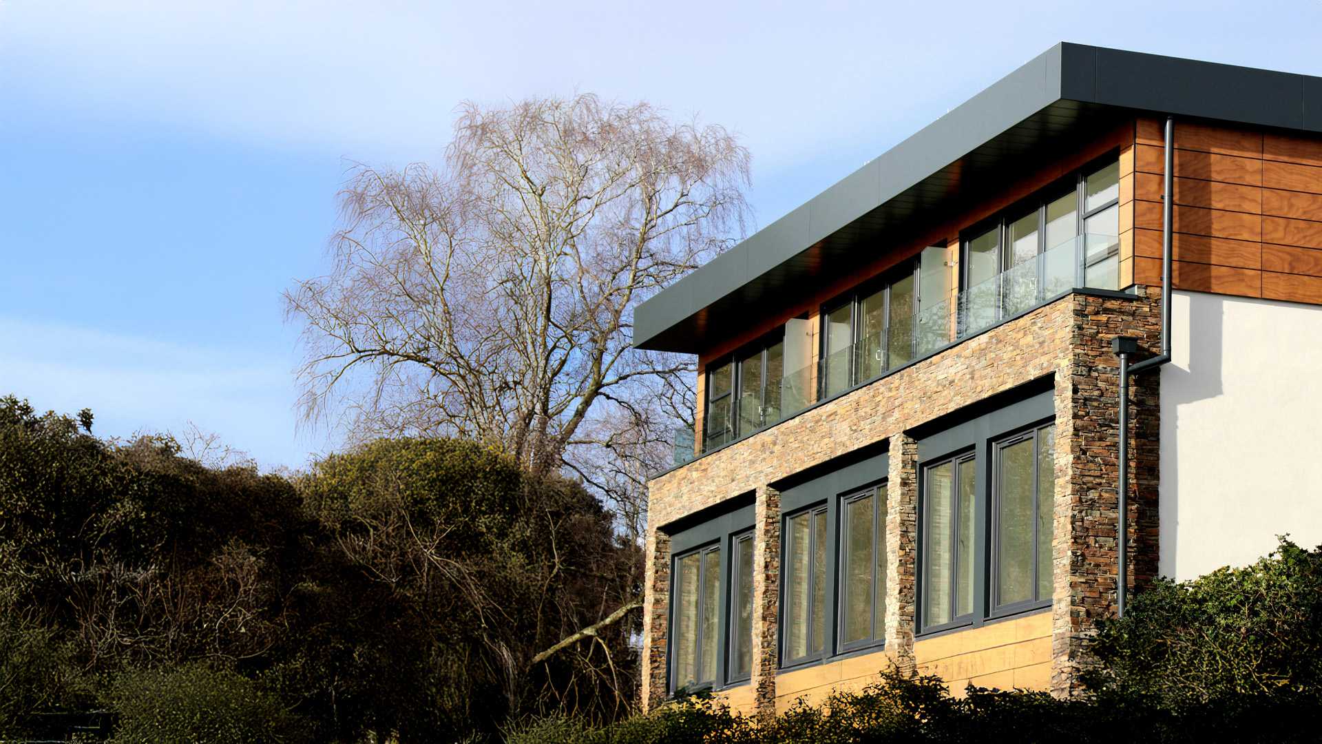 "Modern flat-roof building with large windows, stone and wood exterior, surrounded by trees and under a clear blue sky.