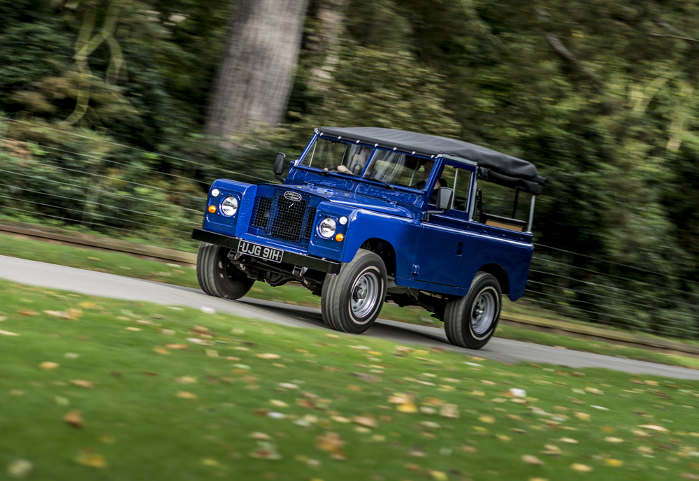 A classic blue Land Rover Series IIA with a black soft top drives on a tree-lined road, surrounded by green grass and scattered autumn leaves—perfectly protected with Agreed Value Car Insurance.