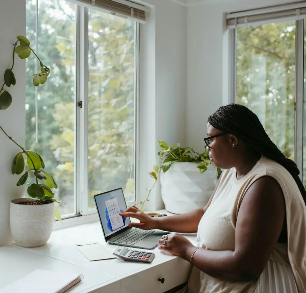 A woman sits at a white desk by a window, using a tablet and calculator—her workspace for managing her home business insurance. Plants and a notebook are on the desk, with greenery visible outside through the windows.