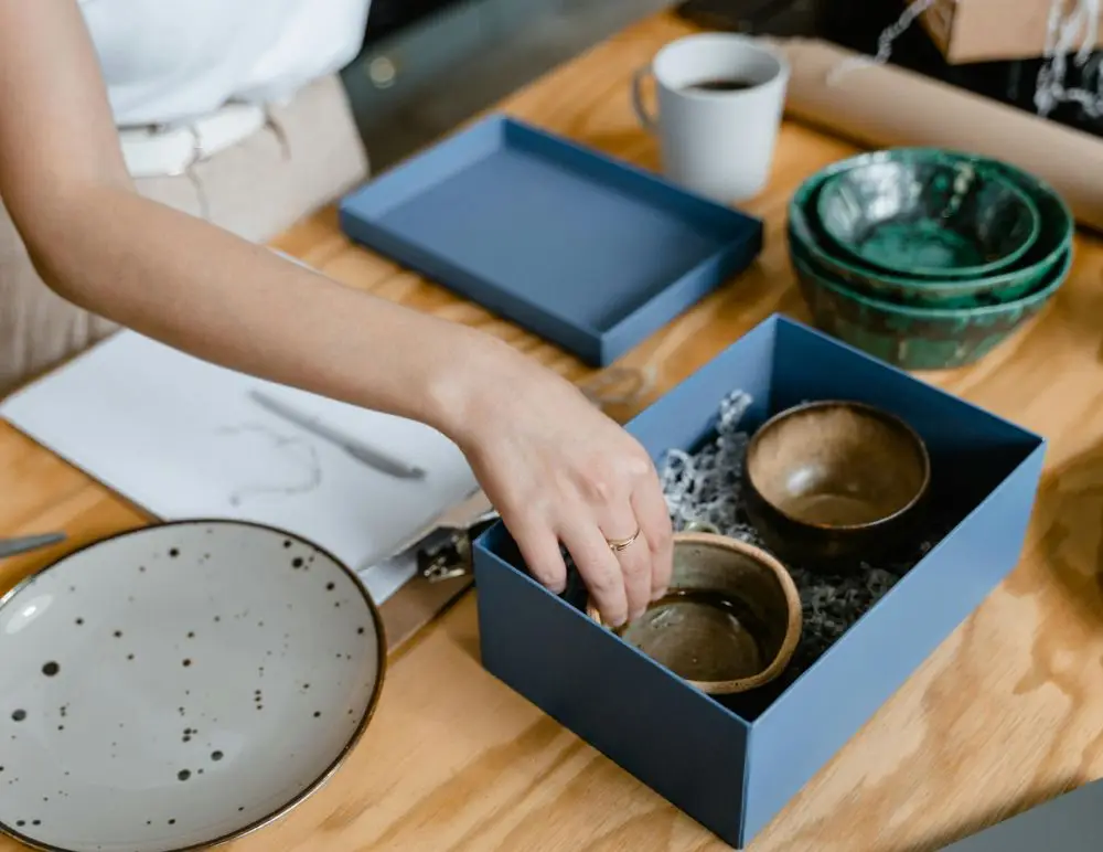 A person packs two small brown bowls into a blue gift box filled with packing material, beside a stack of green bowls, a speckled plate, and a white mug on a wooden table—a thoughtful touch for any home business insurance shipping process.