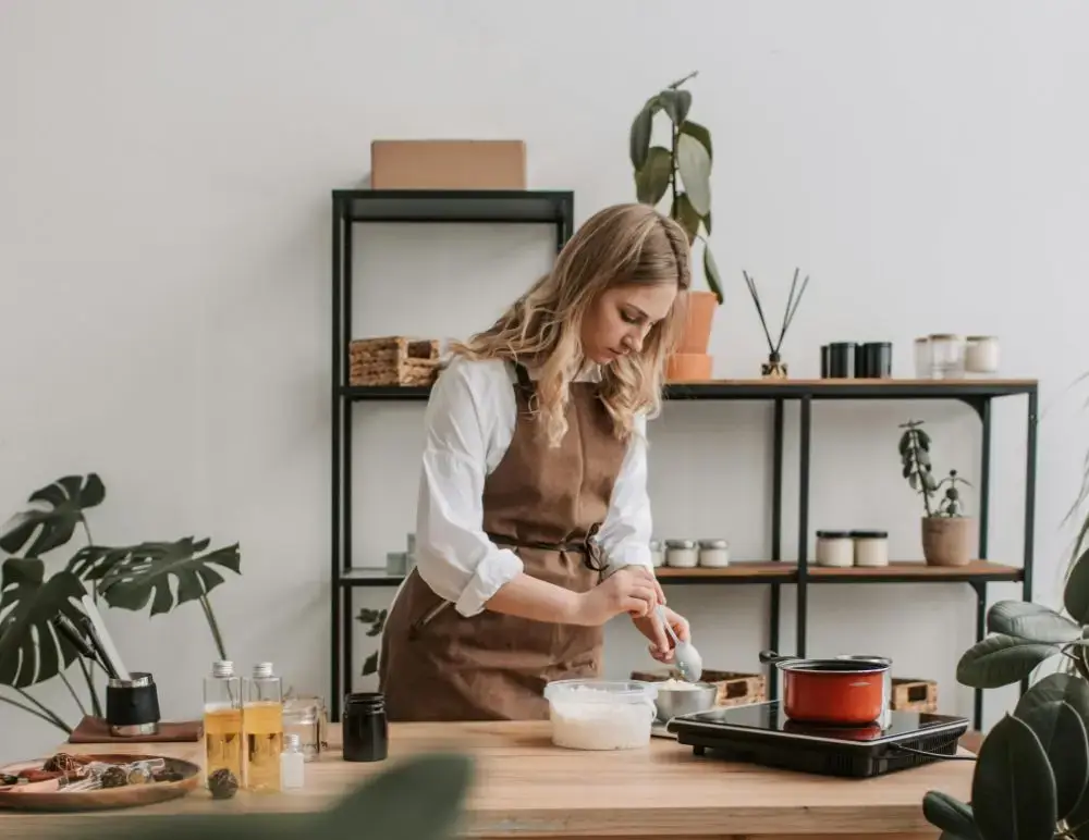 A woman in a brown apron stands at a wooden table, mixing ingredients in a bowl. Bottles, jars, and a red pot sit on a portable hob—an ideal setup for those considering Home Business Insurance whilst running their kitchen business.