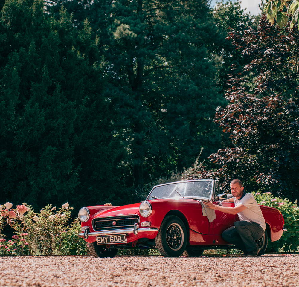 A man crouches beside a classic red convertible car, parked on a gravel drive surrounded by greenery and flowering shrubs on a sunny day—an ideal scene for Agreed Value Car Insurance protection.